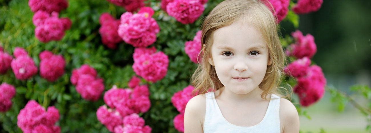 Bambina con capelli rossi in posa davanti a un cespuglio di gerani rosa